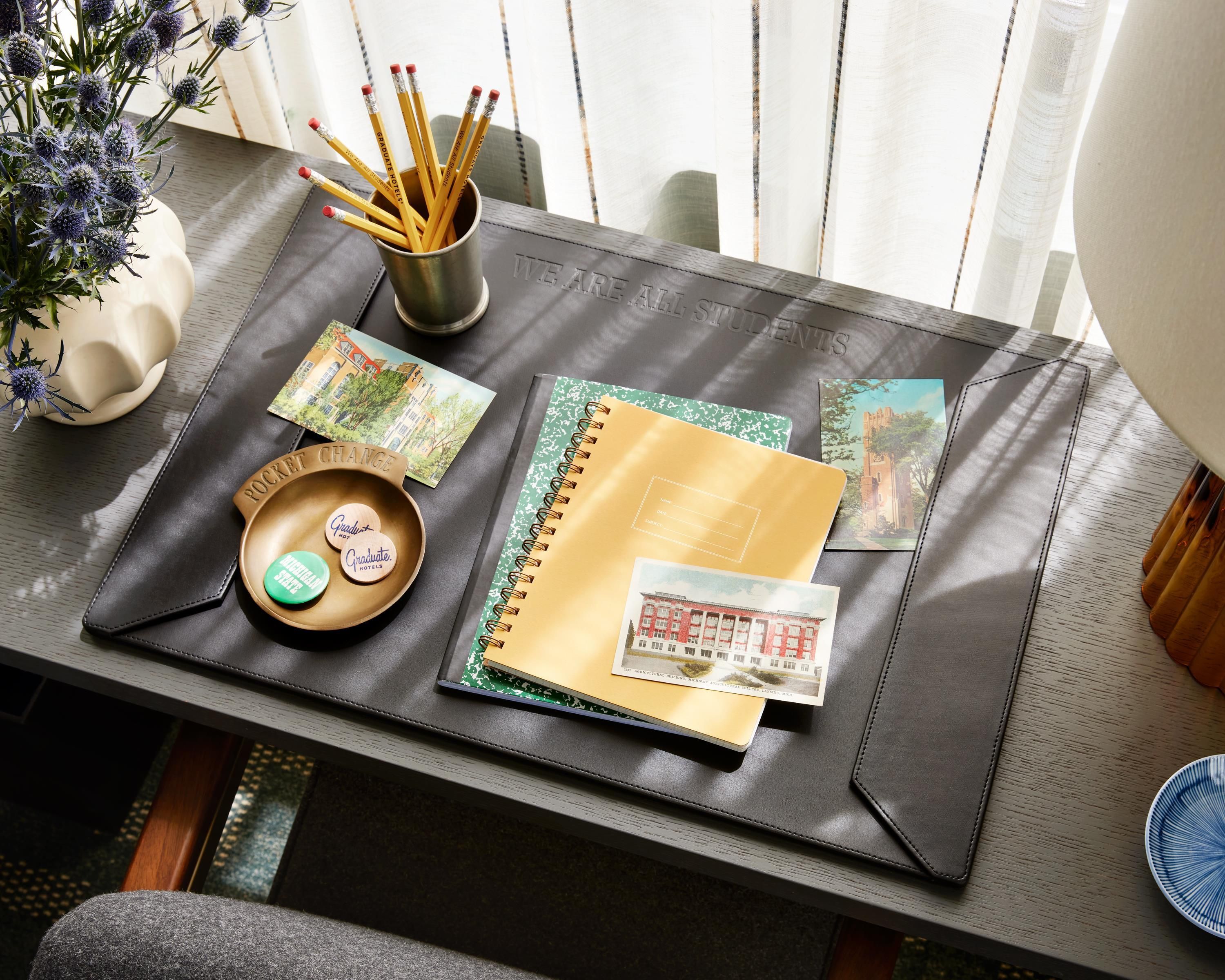 A desk with a leather desk pad, a notebook, a pencil holder with pencils, a small potted plant, and a couple of business cards.