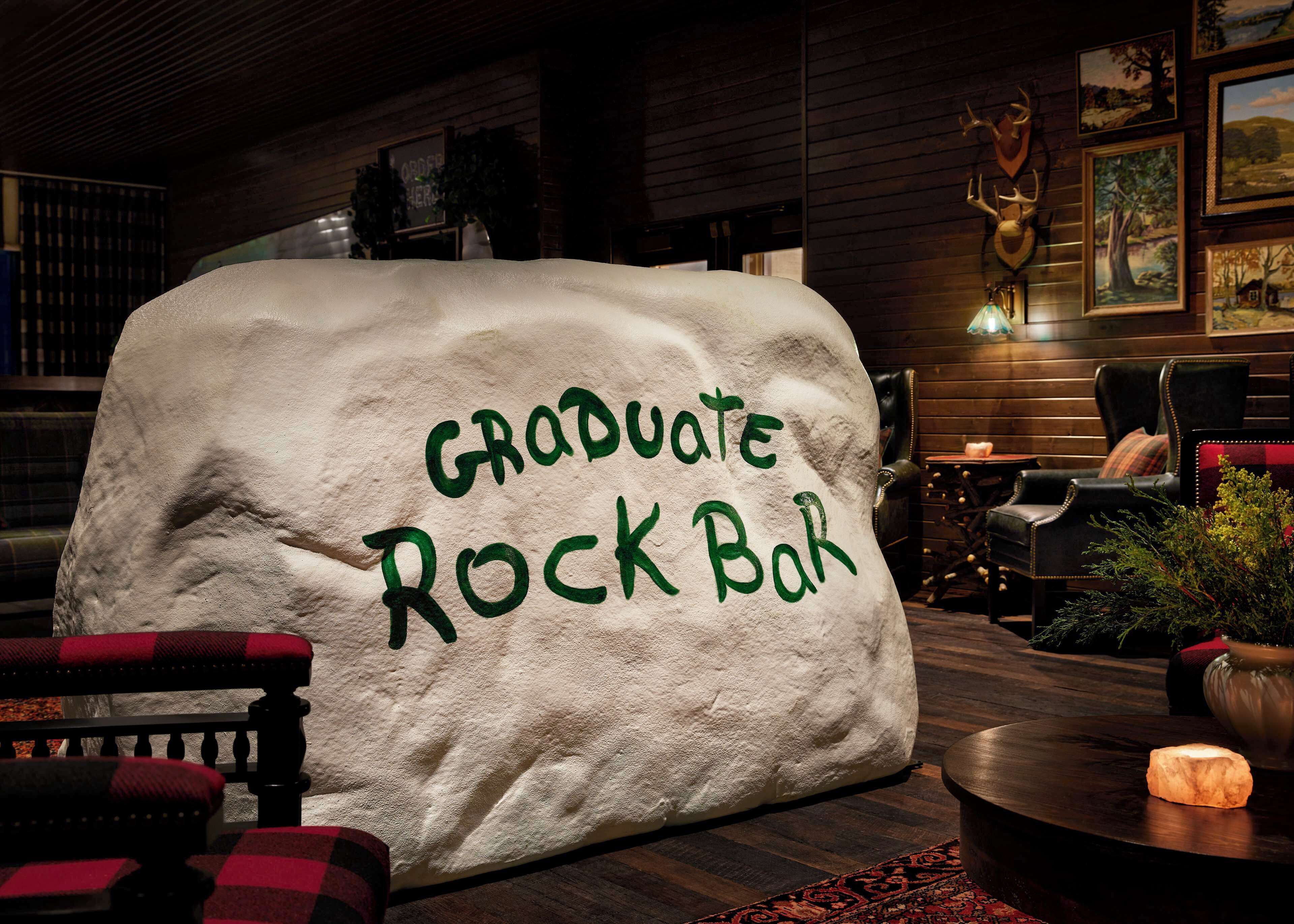A cozy bar area with a large rock centerpiece labeled "Graduate Rock Bar," wooden furniture, and framed artwork on the walls.