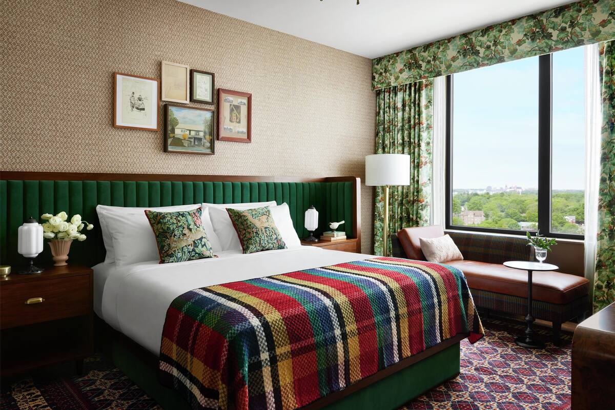 Une chambre d’hôtel avec un grand lit, une tête de lit recouverte d’un tissu vert et ornée de motifs floraux, des rideaux à fleurs et une vue sur la ville.