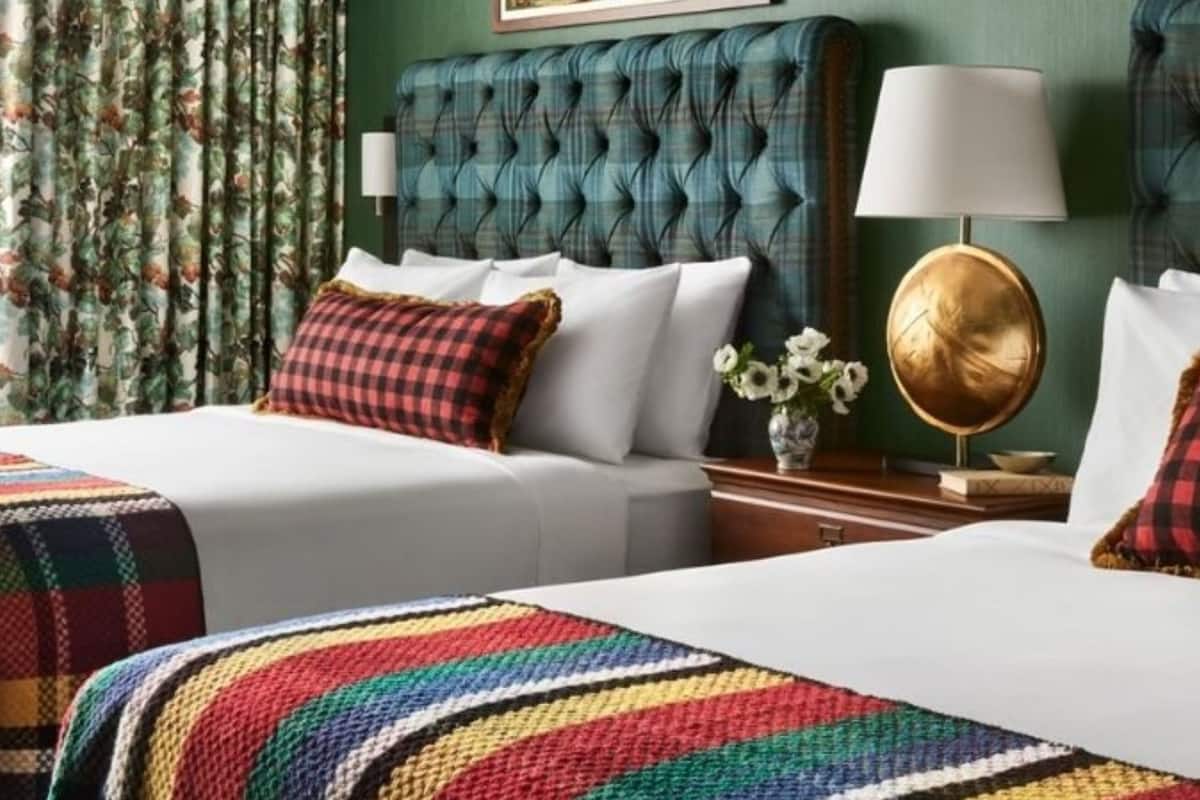 Une chambre d’hôtel avec une tête de lit capitonnée, un lit blanc recouvert d’une couverture colorée, une table de chevet avec une lampe et des rideaux à motifs floraux.