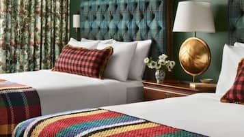 A hotel room with a tufted headboard, a white bed with a colorful blanket, a bedside table with a lamp, and floral curtains.