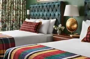 A hotel room with a tufted headboard, a white bed with a colorful blanket, a bedside table with a lamp, and floral curtains.
