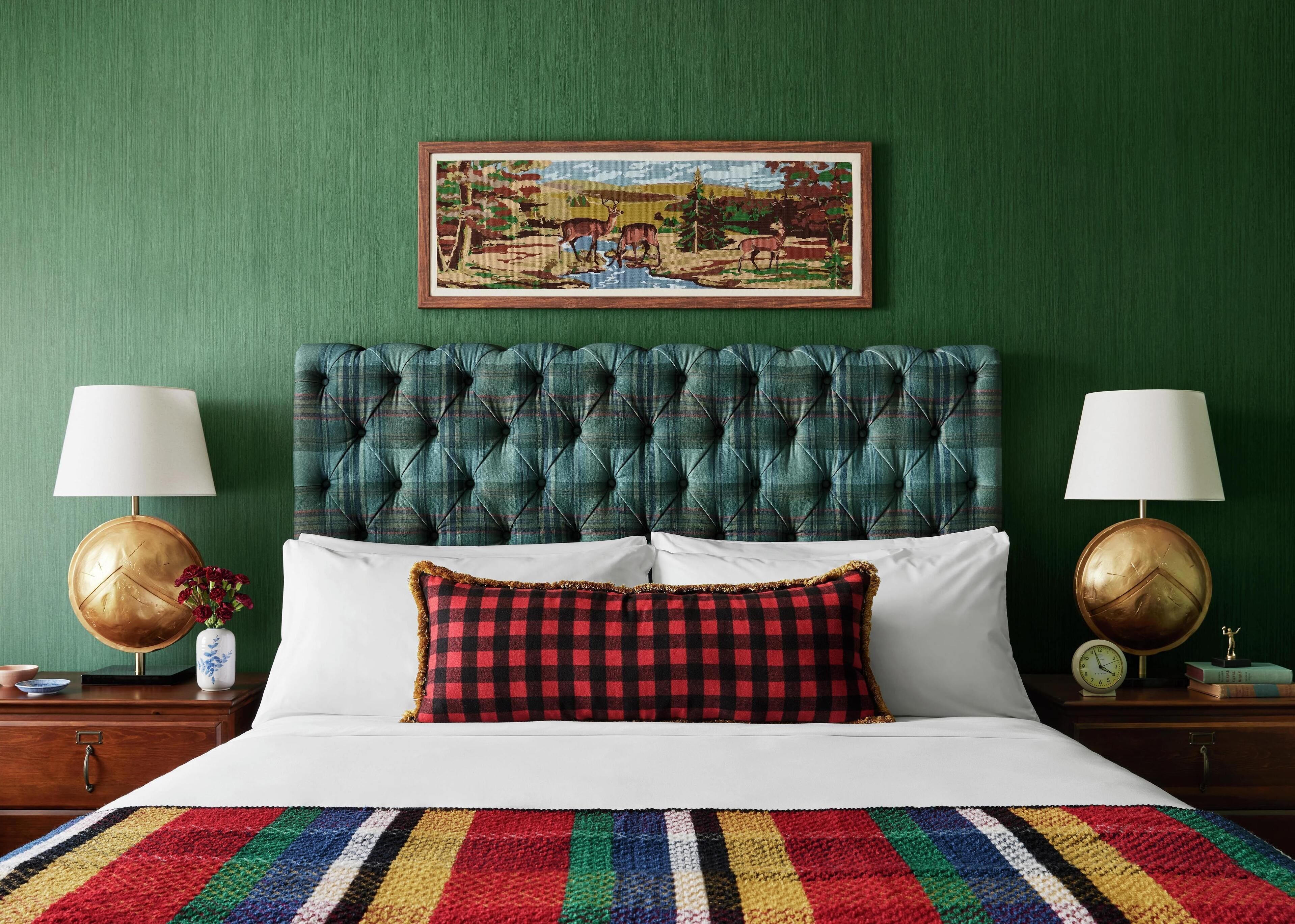 Una cama bien hecha con un edredón a rayas de colores, una almohada escocesa y un cabecero capitonado. Hay dos mesitas de noche con lámparas y un cuadro enmarcado sobre la cama.