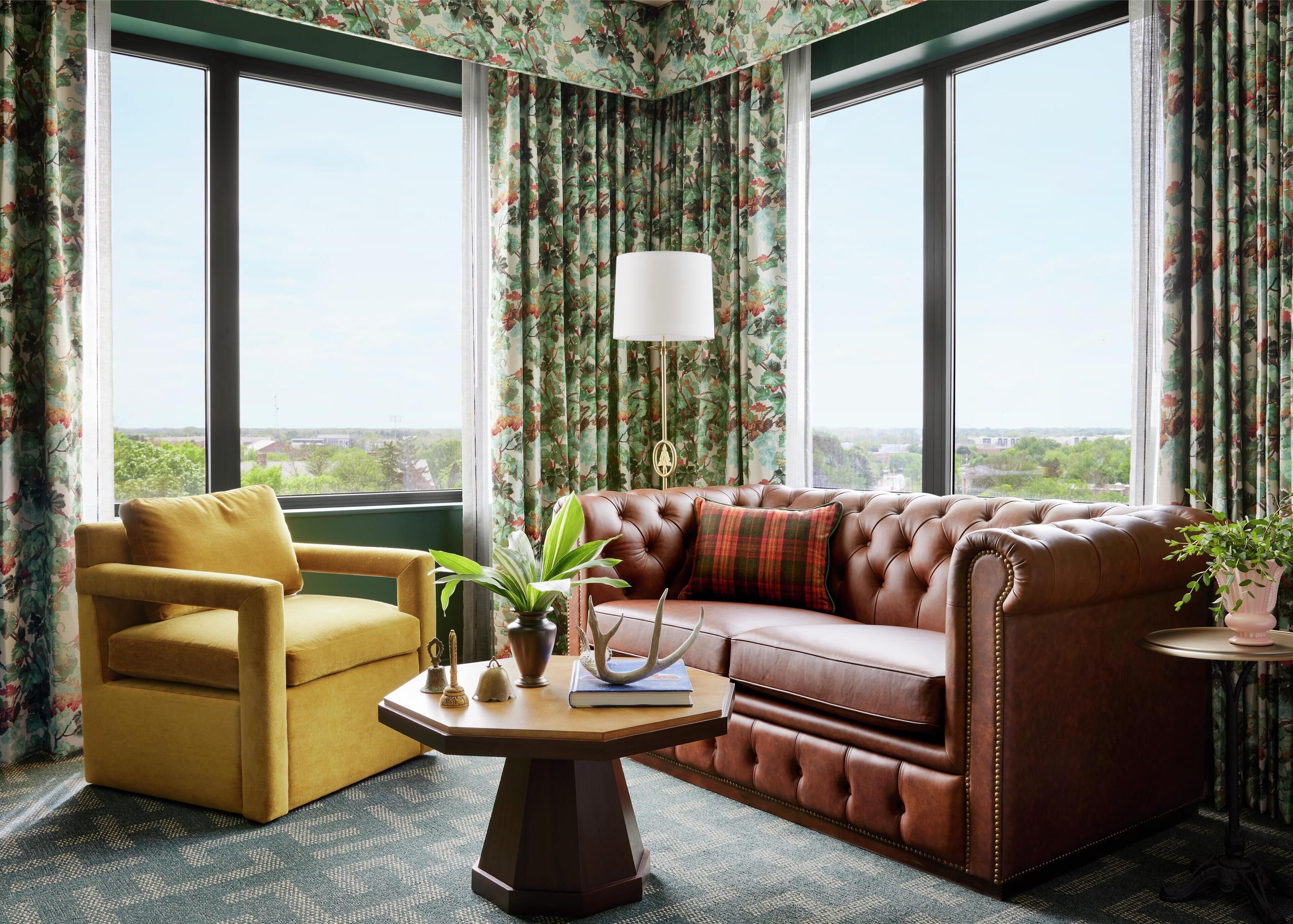 Una habitación con un sofá de cuero, un sillón amarillo, una mesita con una lámpara y cortinas con estampado floral.