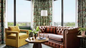Una habitación con un sofá de cuero, un sillón amarillo, una mesita con una lámpara y cortinas con estampado floral.