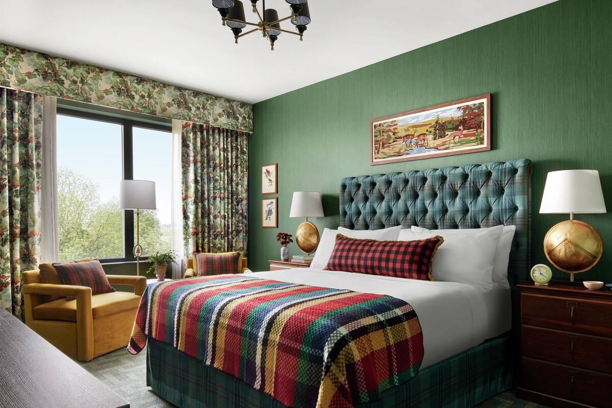 Une chambre d’hôtel avec un grand lit, des murs verts, des rideaux à motifs floraux, une tête de lit capitonnée et un tableau au-dessus du lit.