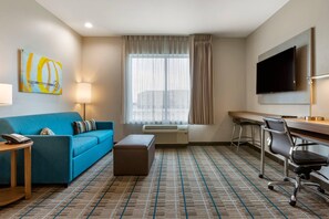 A hotel room with a blue sofa, a small table with a telephone, a TV mounted on the wall, a desk with a chair, and an air conditioning unit. - MainStay Suites Carlsbad South (Carlsbad)