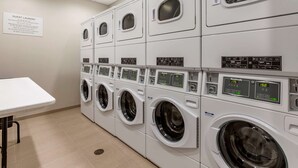 A row of stacked front-loading washing machines with control panels, a guest laundry sign, and a white table. - MainStay Suites Carlsbad South (Carlsbad)