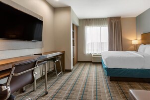 Habitación de hotel con televisión de pantalla plana, un escritorio con silla, cama con detalles azules y una ventana con cortinas.