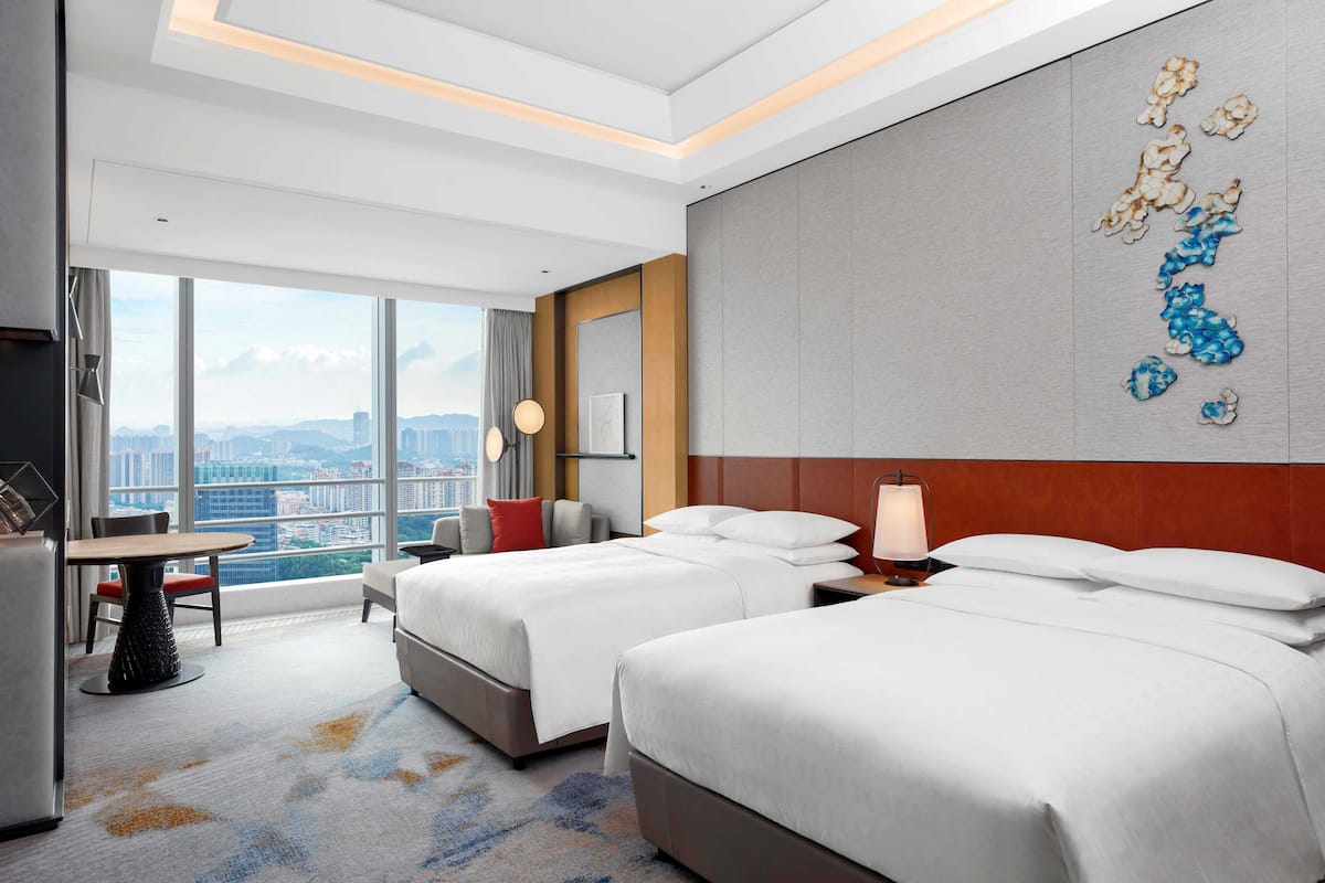 A hotel room with two beds, a dining table, and a large window with a city view.