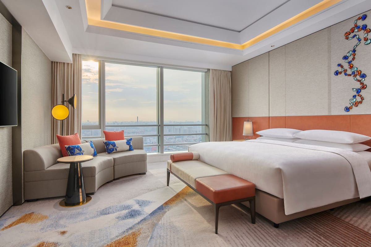 A modern hotel room with a large bed, a sofa, a small table, and a view of the city.