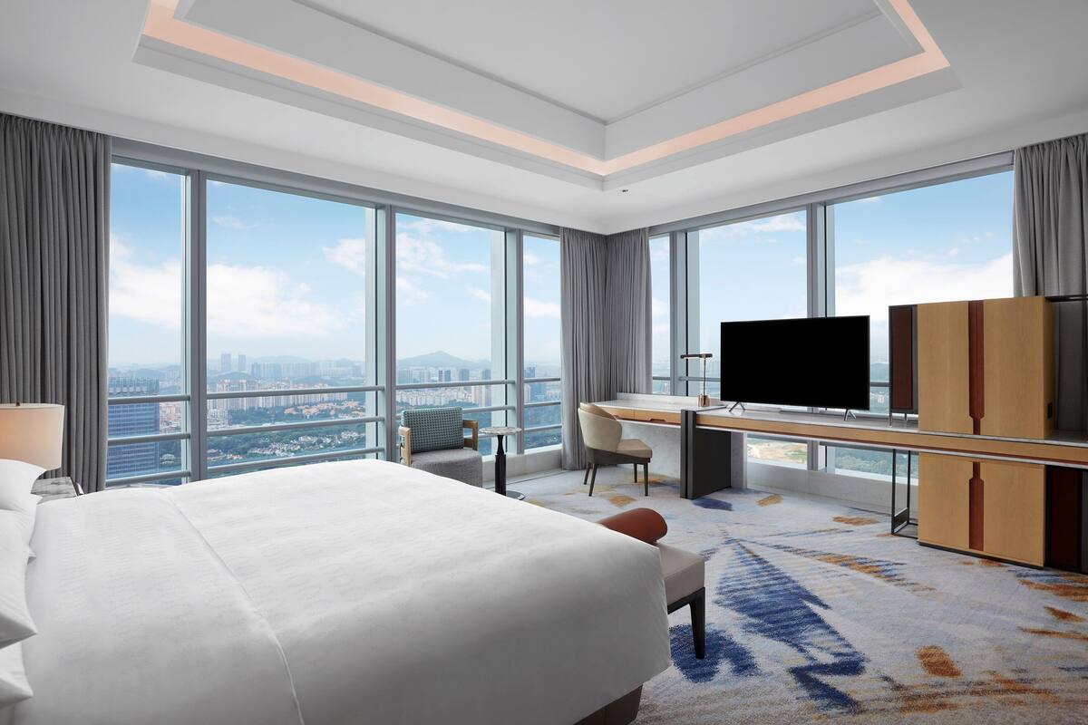 A modern hotel room with a large bed, a desk with a flat-screen TV, and a view of the cityscape through large windows.
