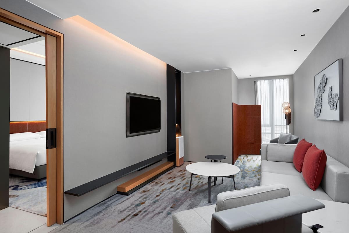 A modern hotel room with a flat-screen TV, a sofa, a small table, and a large window with curtains.
