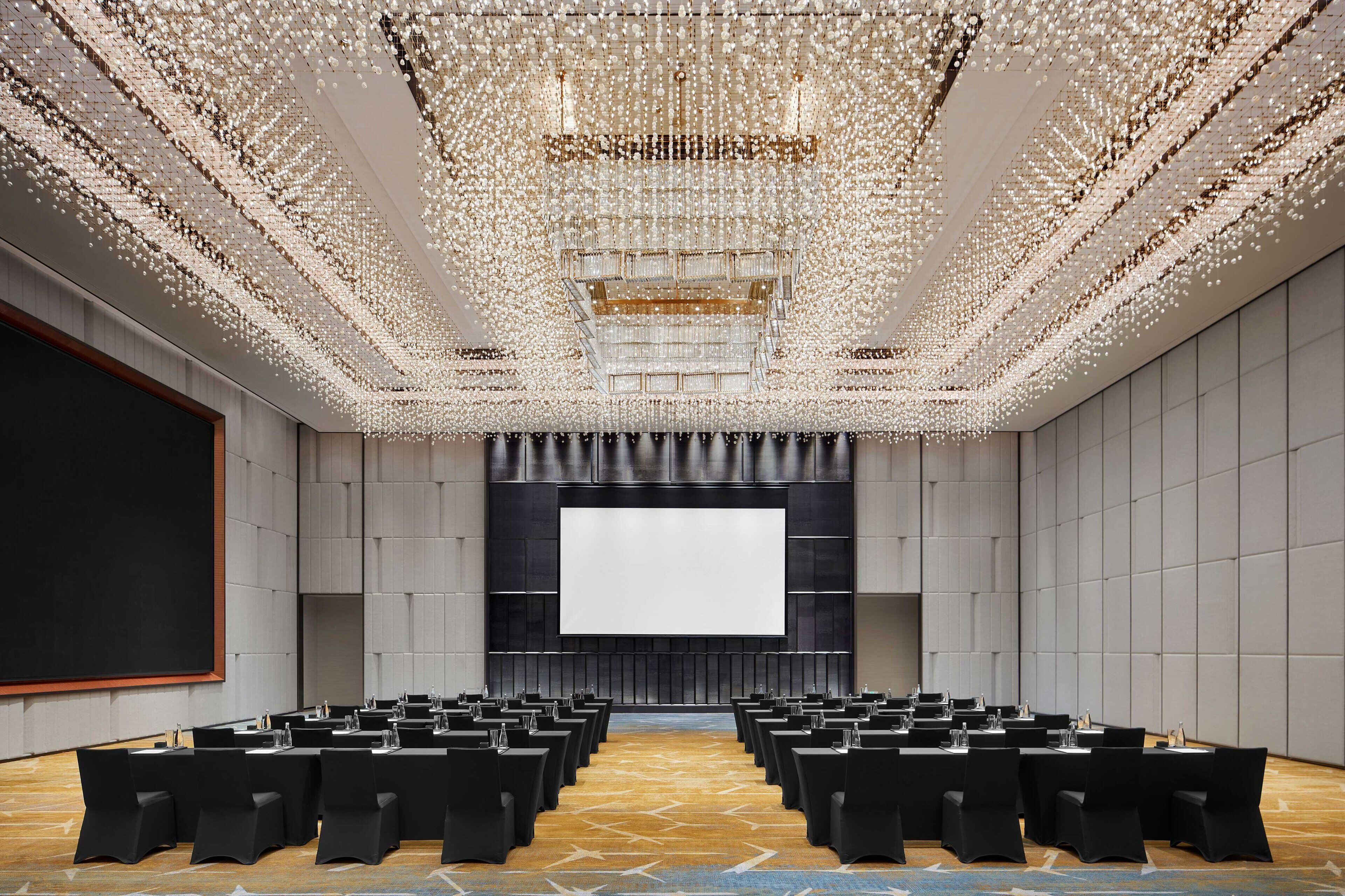 A conference room with rows of tables and chairs, a large screen, and a ceiling with a decorative light fixture.