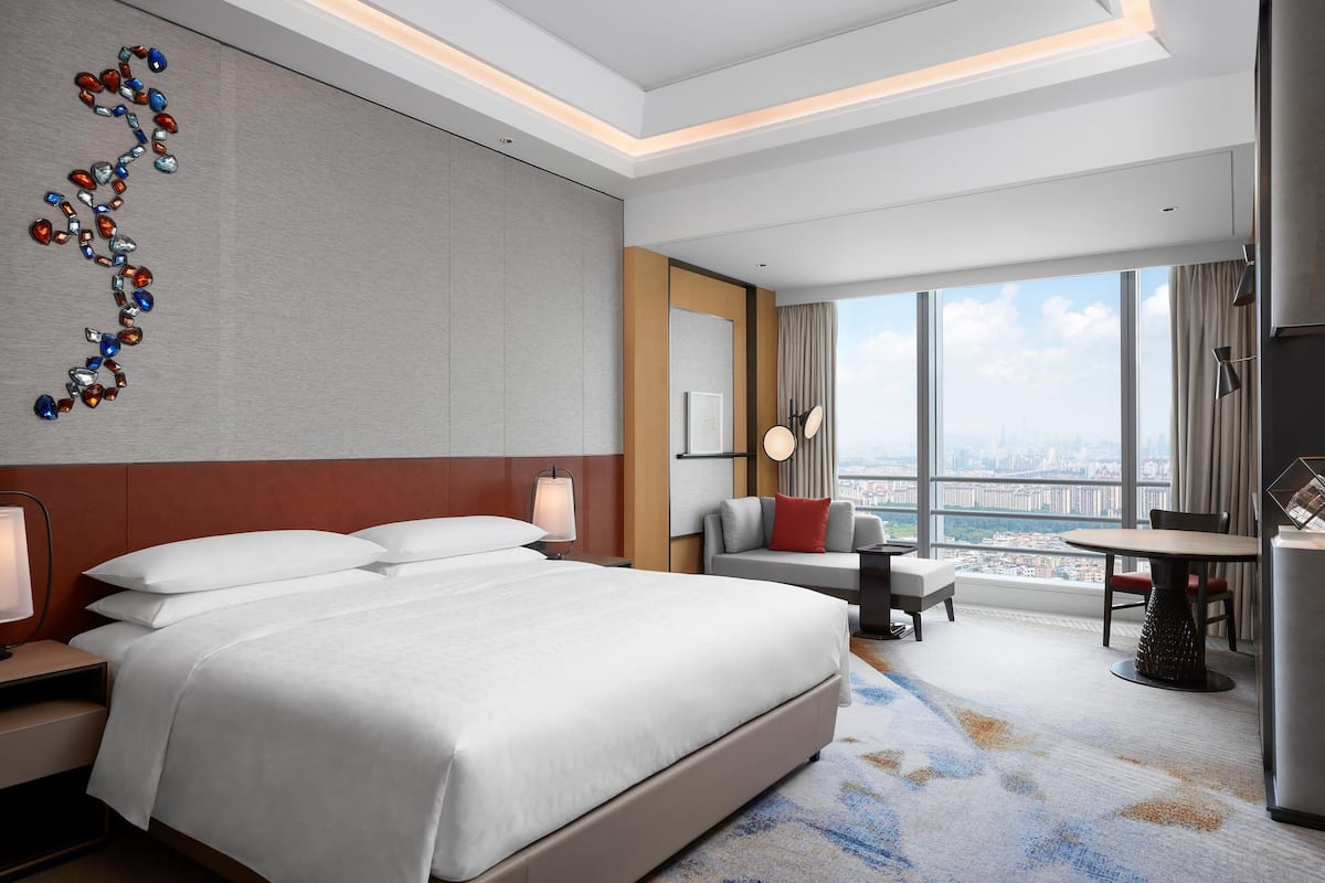 A modern hotel room with a large bed, a desk, a chair, and a view of the city through large windows.