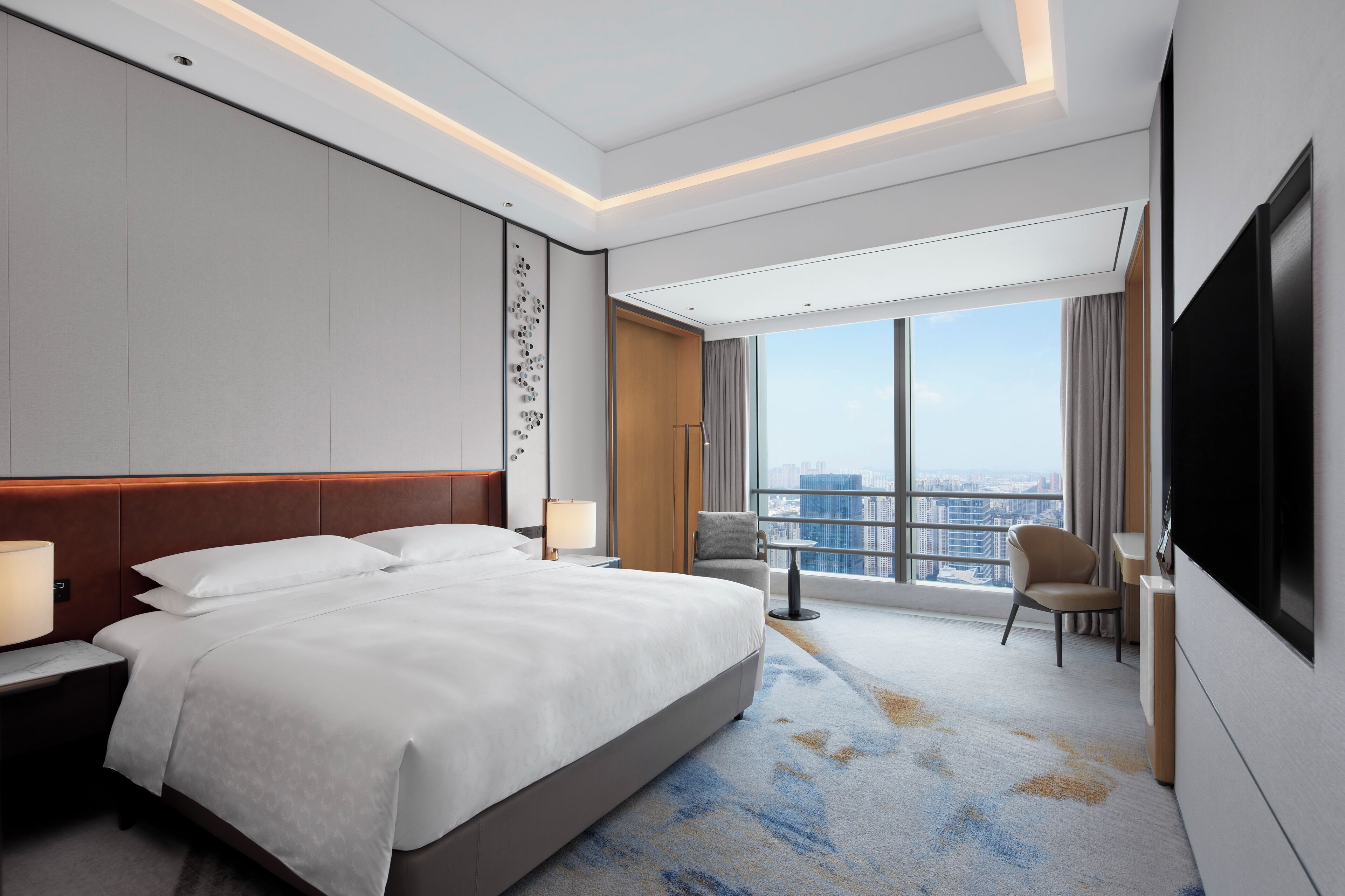 A modern hotel room with a large bed, a flat-screen TV, a desk, and a view of the city.