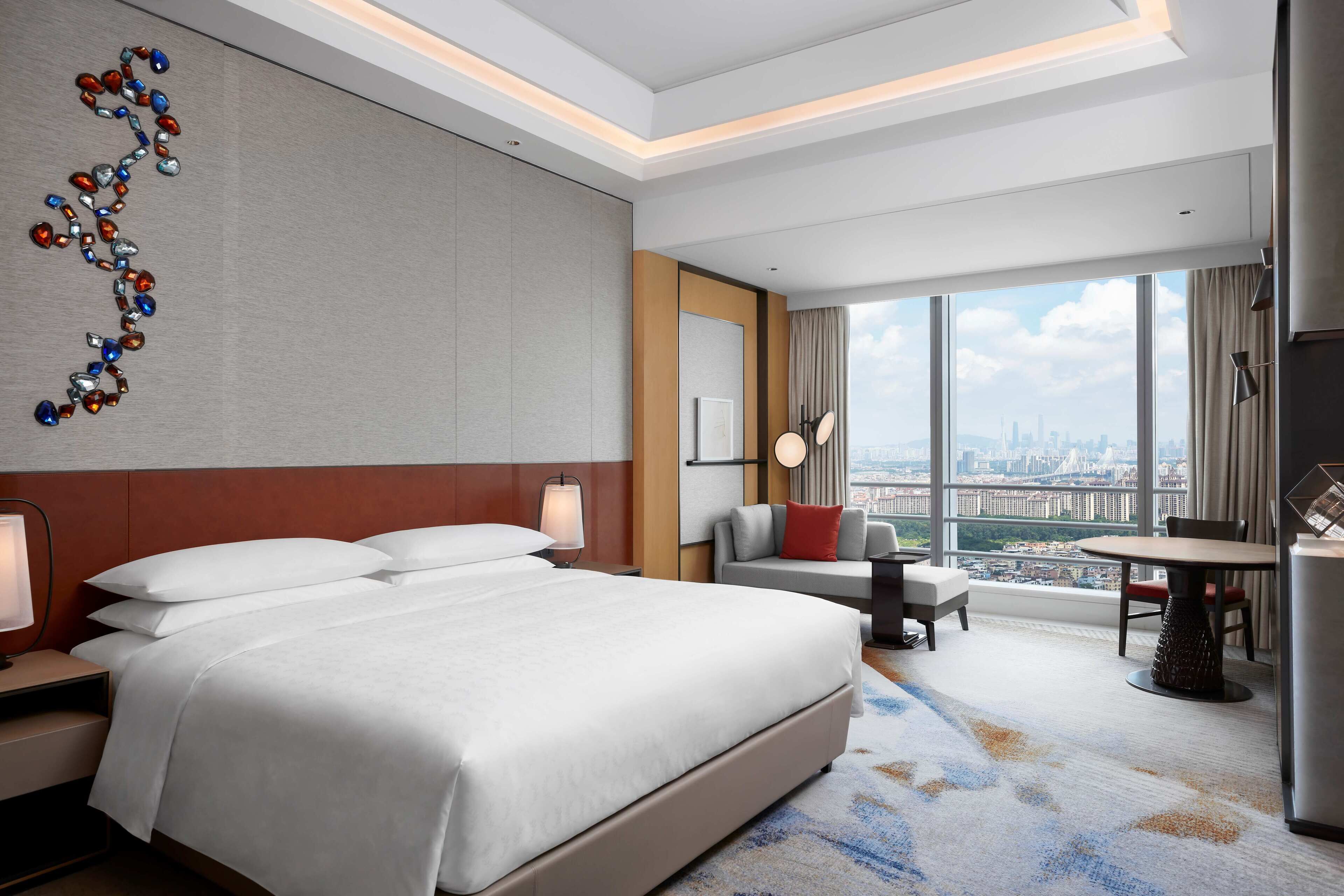 A modern hotel room with a large bed, a desk, a chair, and a view of the city through large windows.