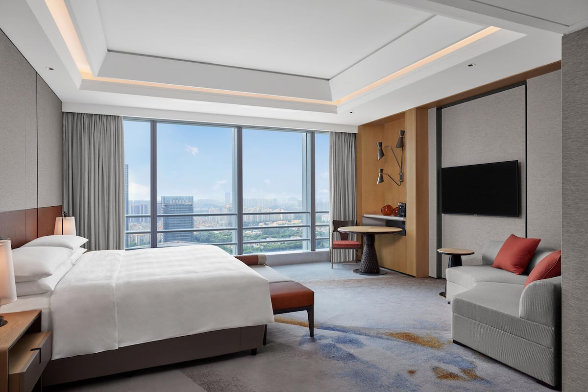 A modern hotel room with a large bed, a sofa, a TV, and a city view.