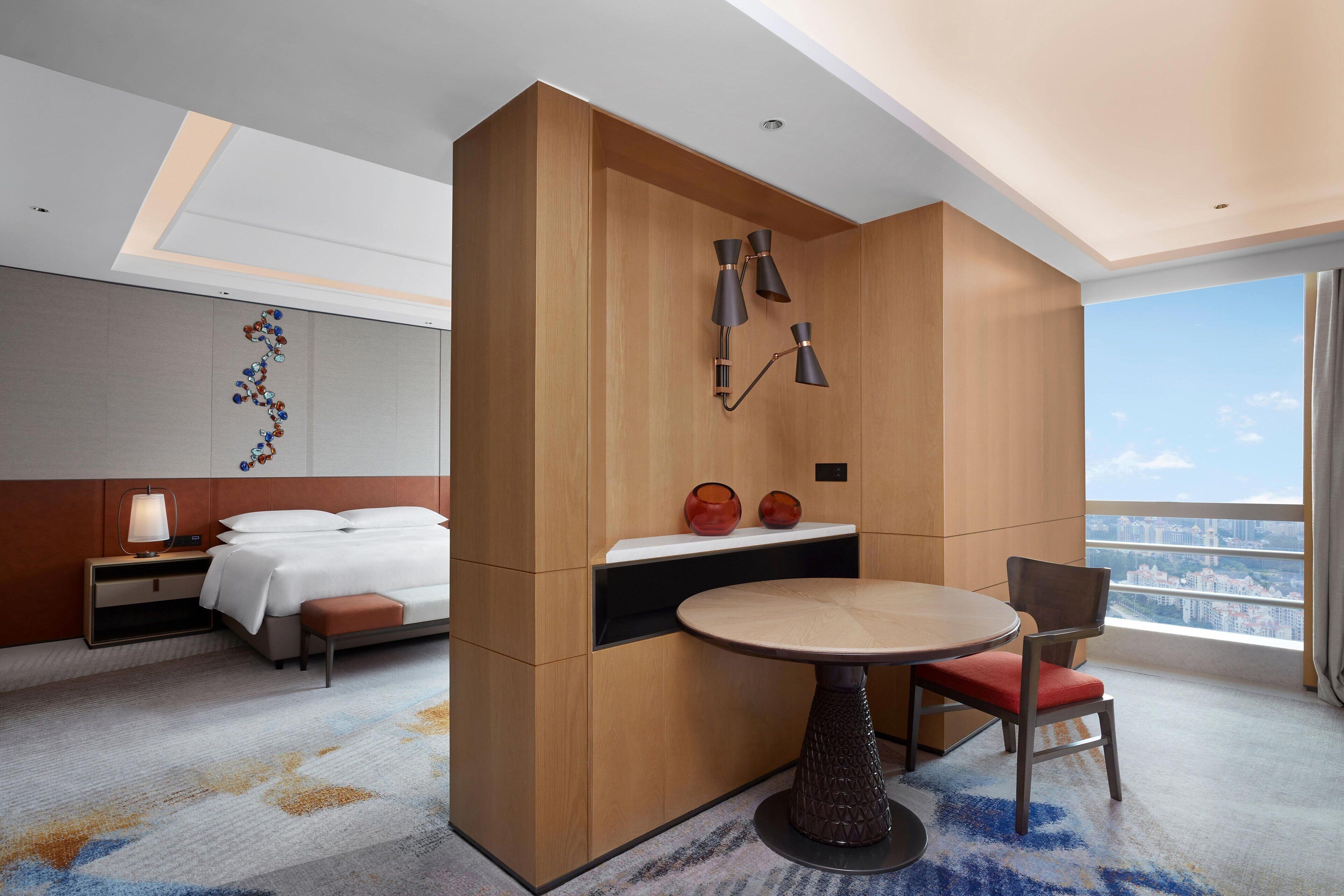 Ein modernes Hotelzimmer mit Bett, Nachttisch, Lampe und einem runden Tisch mit Stuhl. Es gibt ein großes Fenster mit Blick auf die Stadt.