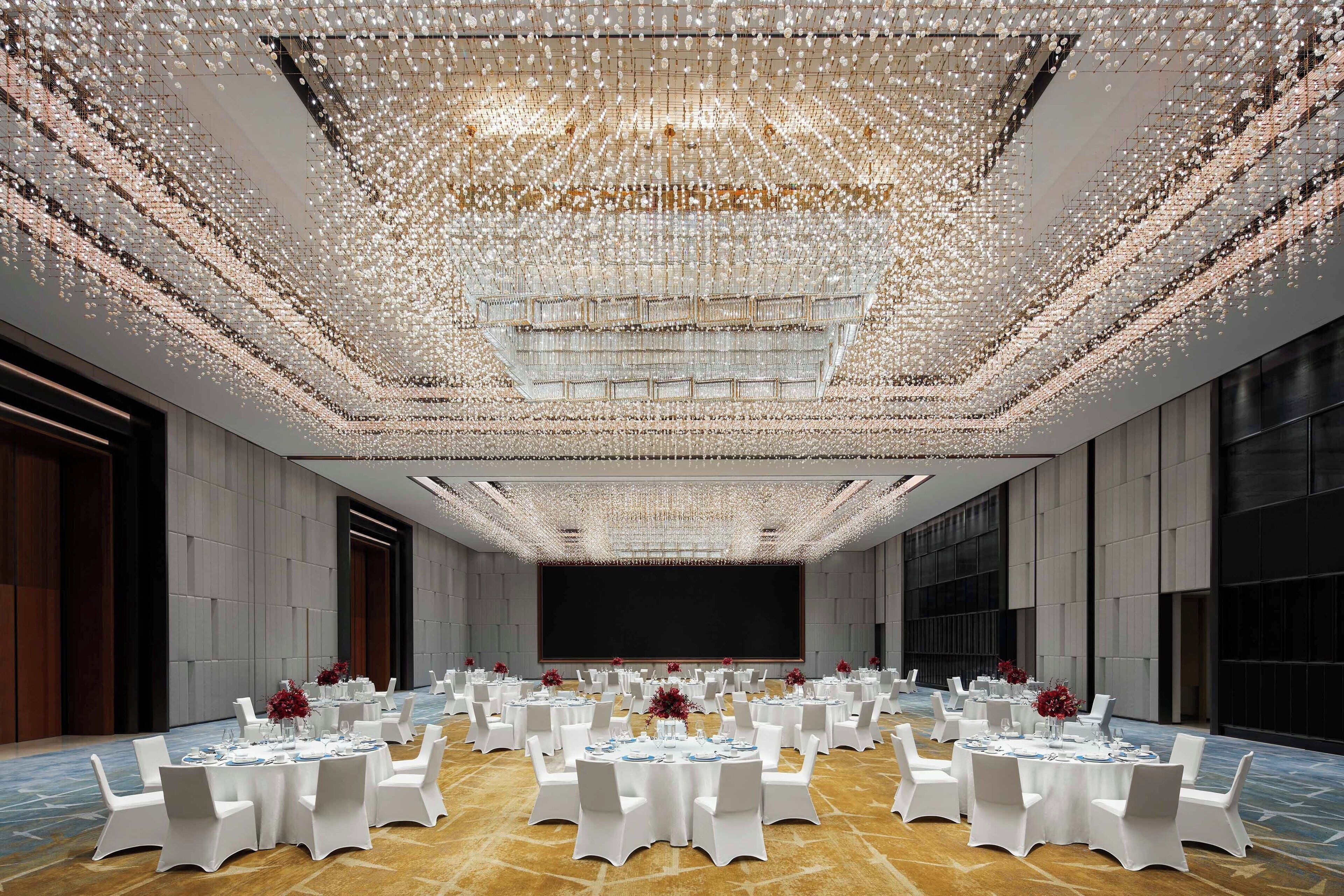 A spacious banquet hall with round tables set for dining, a large screen at the front, and a ceiling adorned with hanging lights.