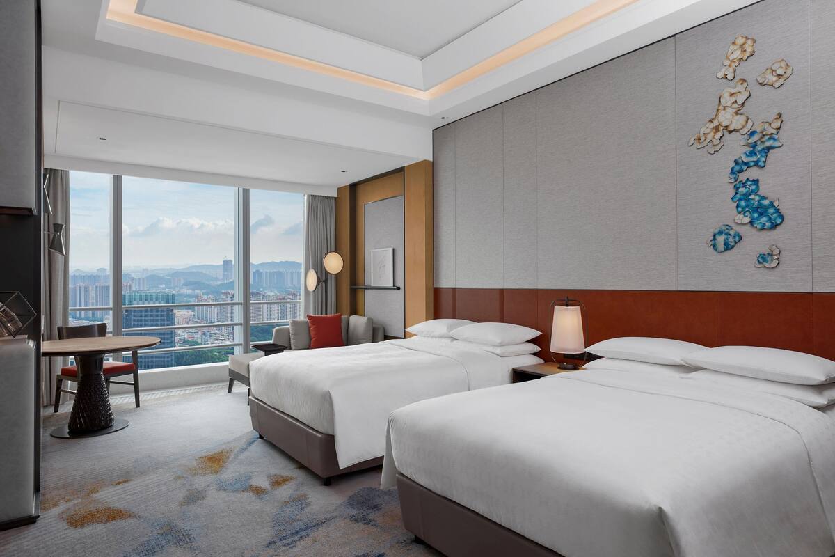 A hotel room with two beds, a dining table, and a large window offering a city view.