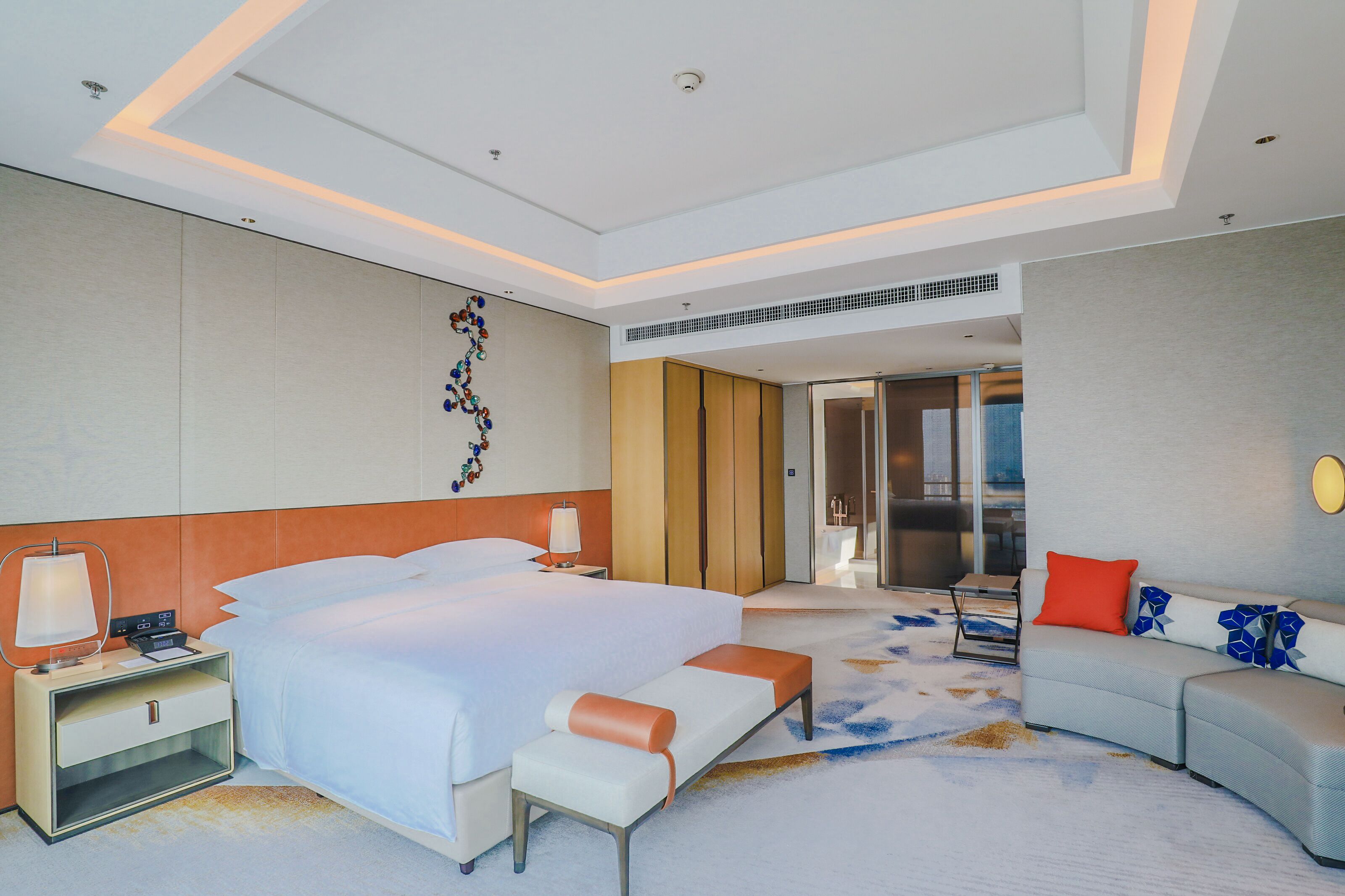 A modern hotel room with a large bed, a bedside table with a lamp, a sofa with a red pillow, and a decorative wall piece.