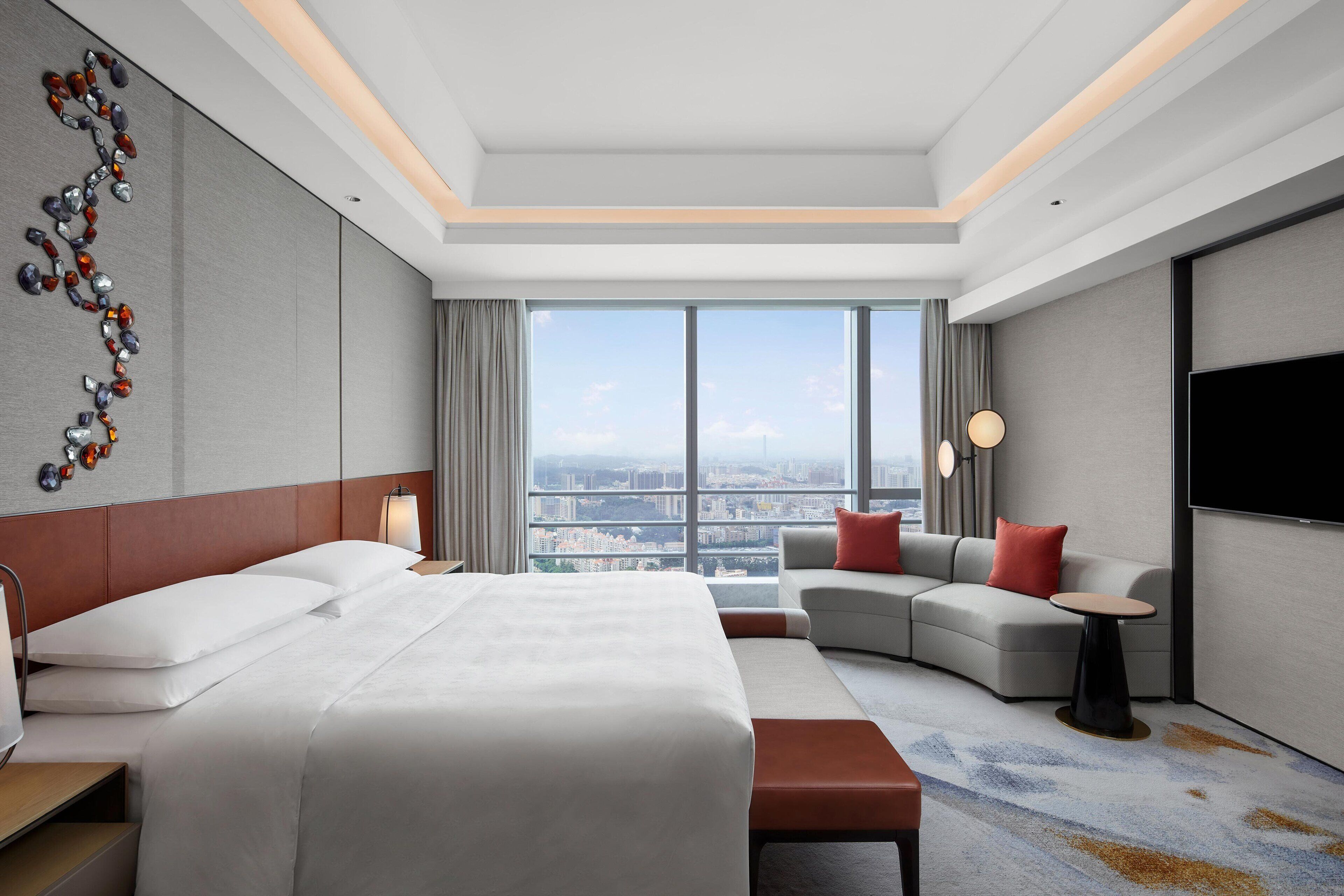 A modern hotel room with a large bed, a sofa, a small table, and a TV. The room has a city view through large windows.