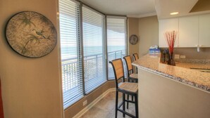 A kitchen with a granite countertop, white cabinets, and a view of the ocean through large windows with blinds.