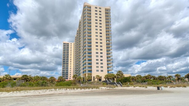 A high-rise hotel with multiple balconies, a sandy beachfront, and palm trees.