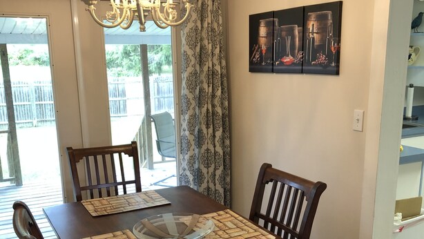 Comedor con mesa de madera, cuatro sillas, una lámpara de araña y vista al exterior a través de un ventanal corredera.