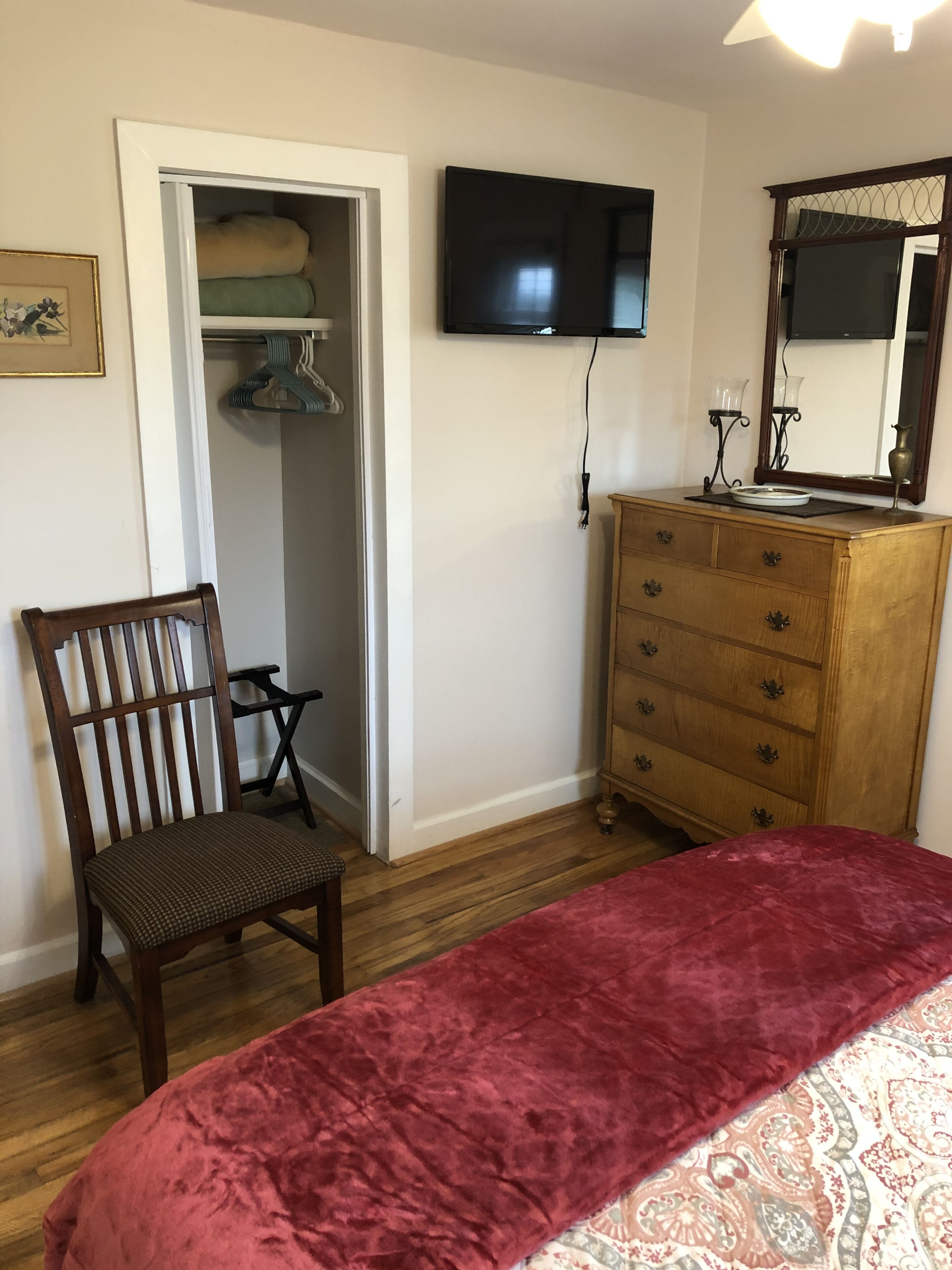 A bedroom with a wooden dresser, a chair, a bed with a red blanket, a television on the wall, and a closet with hanging clothes.