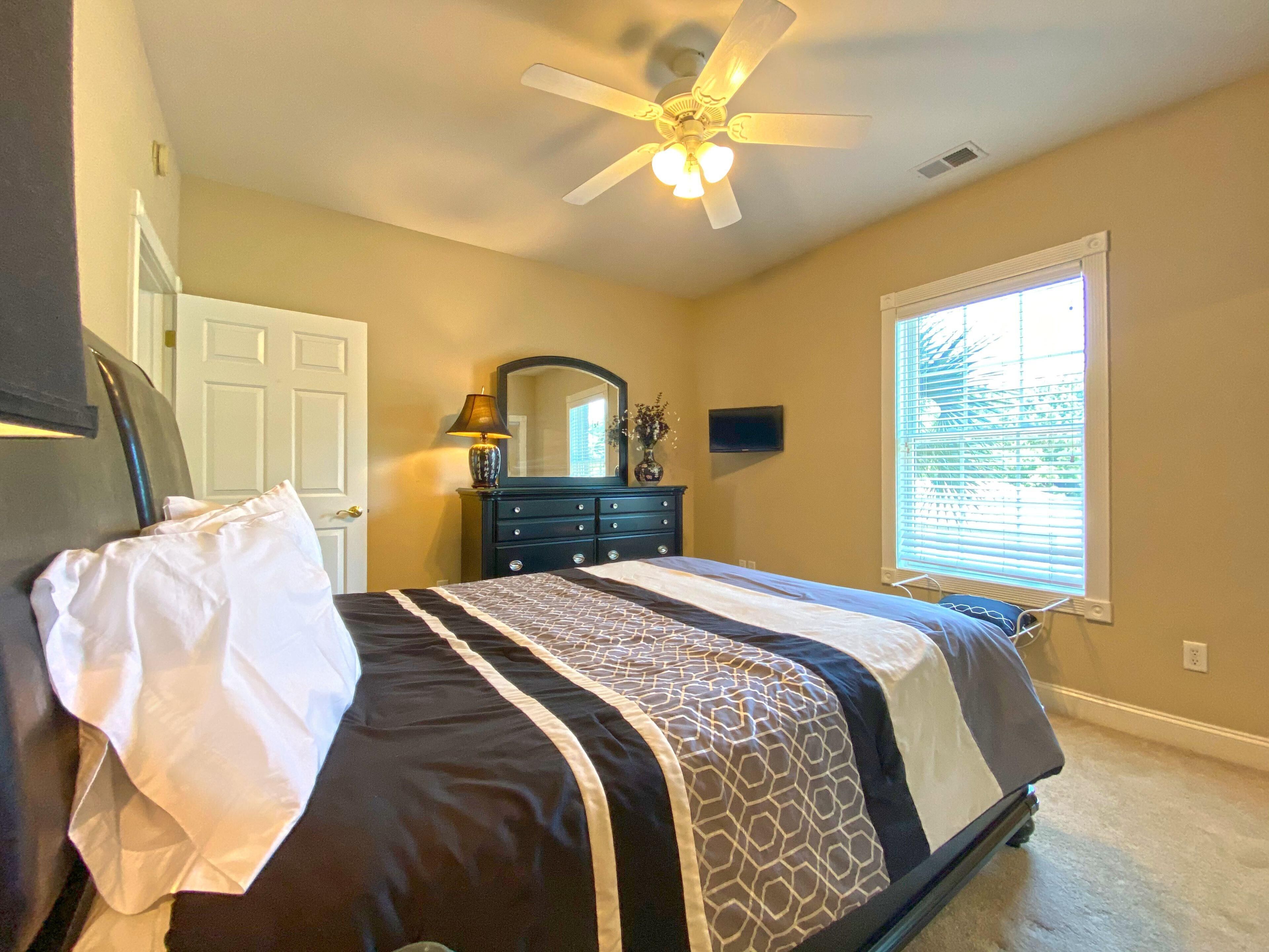 A bedroom with a bed, a ceiling fan, a dresser, a mirror, a window with blinds, and a door.