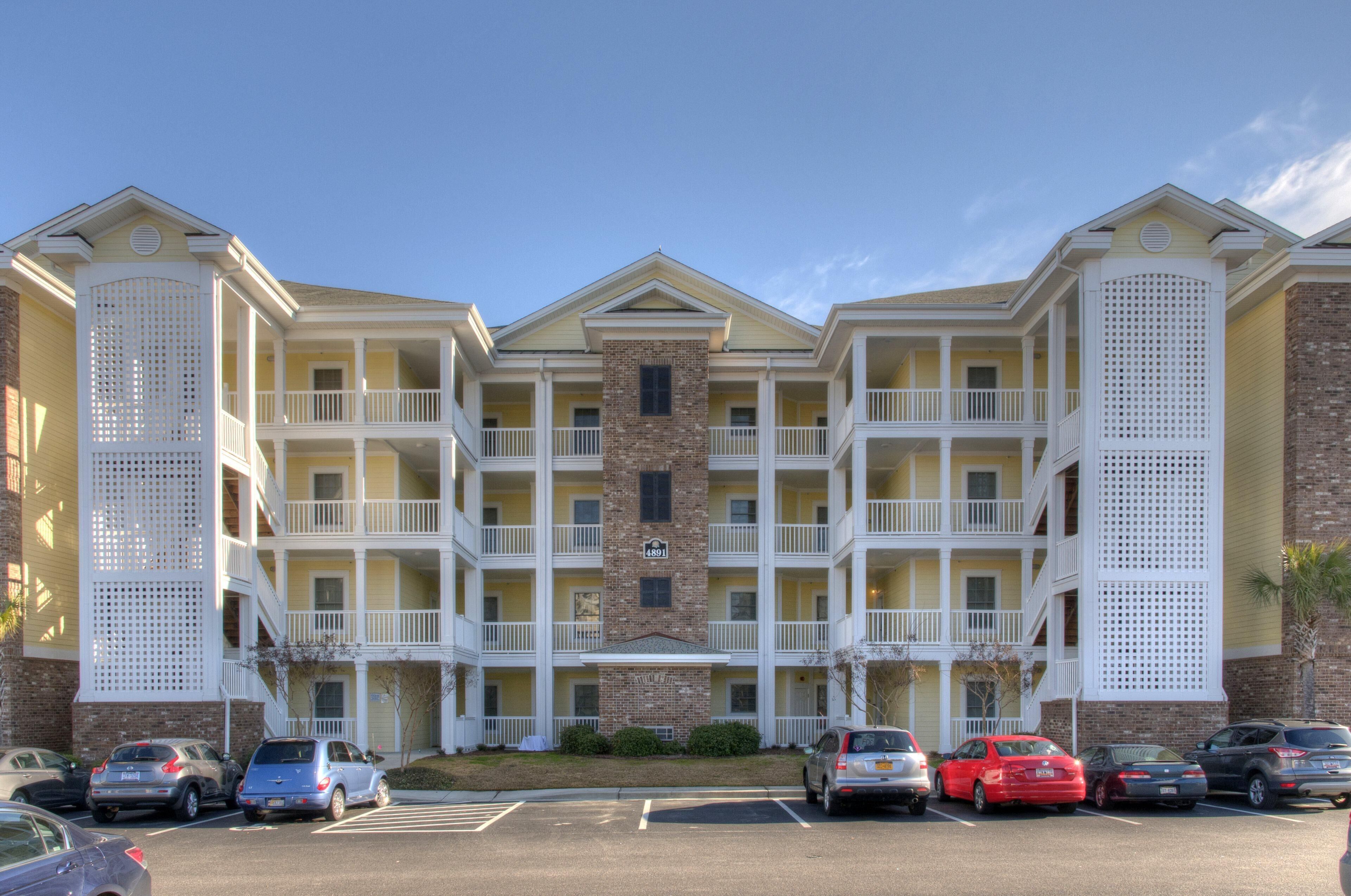A multi-story hotel with a central brick tower, balconies, and a parking lot with several cars.