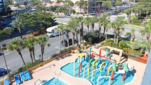 A swimming pool with a slide and a small water play area, surrounded by palm trees and a parking lot. - Oceanview Villa With NEW Furniture! (Myrtle Beach)