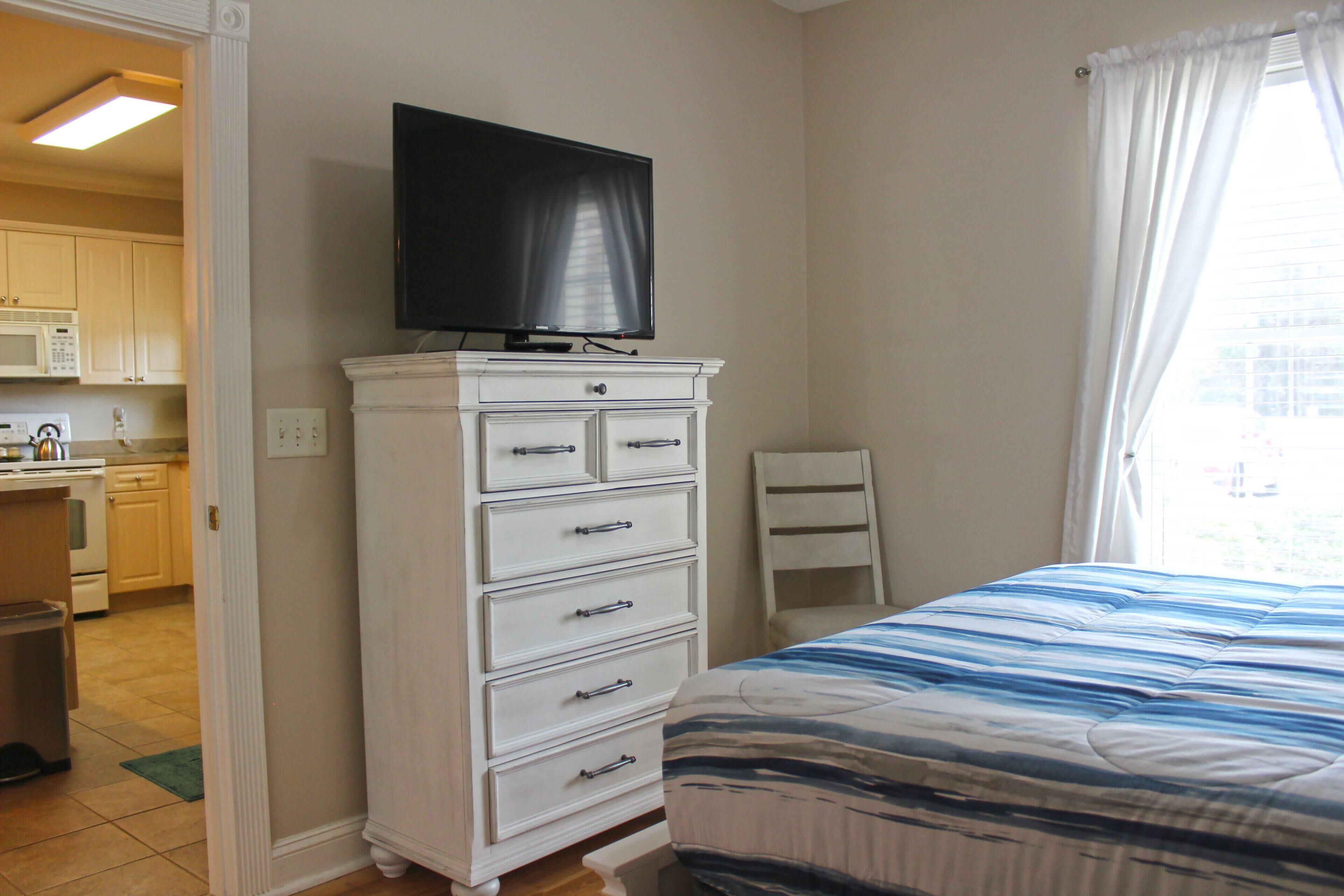 A bedroom with a bed, a dresser with a television on top, a chair, and a view of the outside through a window.