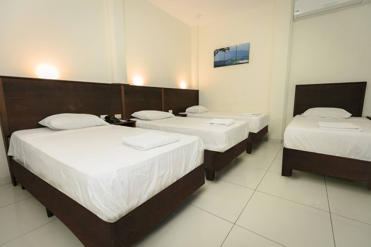 Quarto de hotel com três camas de solteiro, cada uma com cabeceira de madeira e roupa de cama branca. Há um ar condicionado fixado na parede e um pequeno quadro sobre as camas.