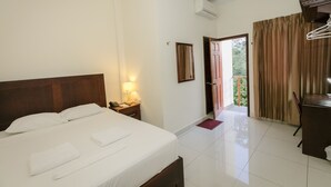 A hotel room with a neatly made bed, a bedside table with a lamp, a desk with a chair, and a view of the outdoors through a door.