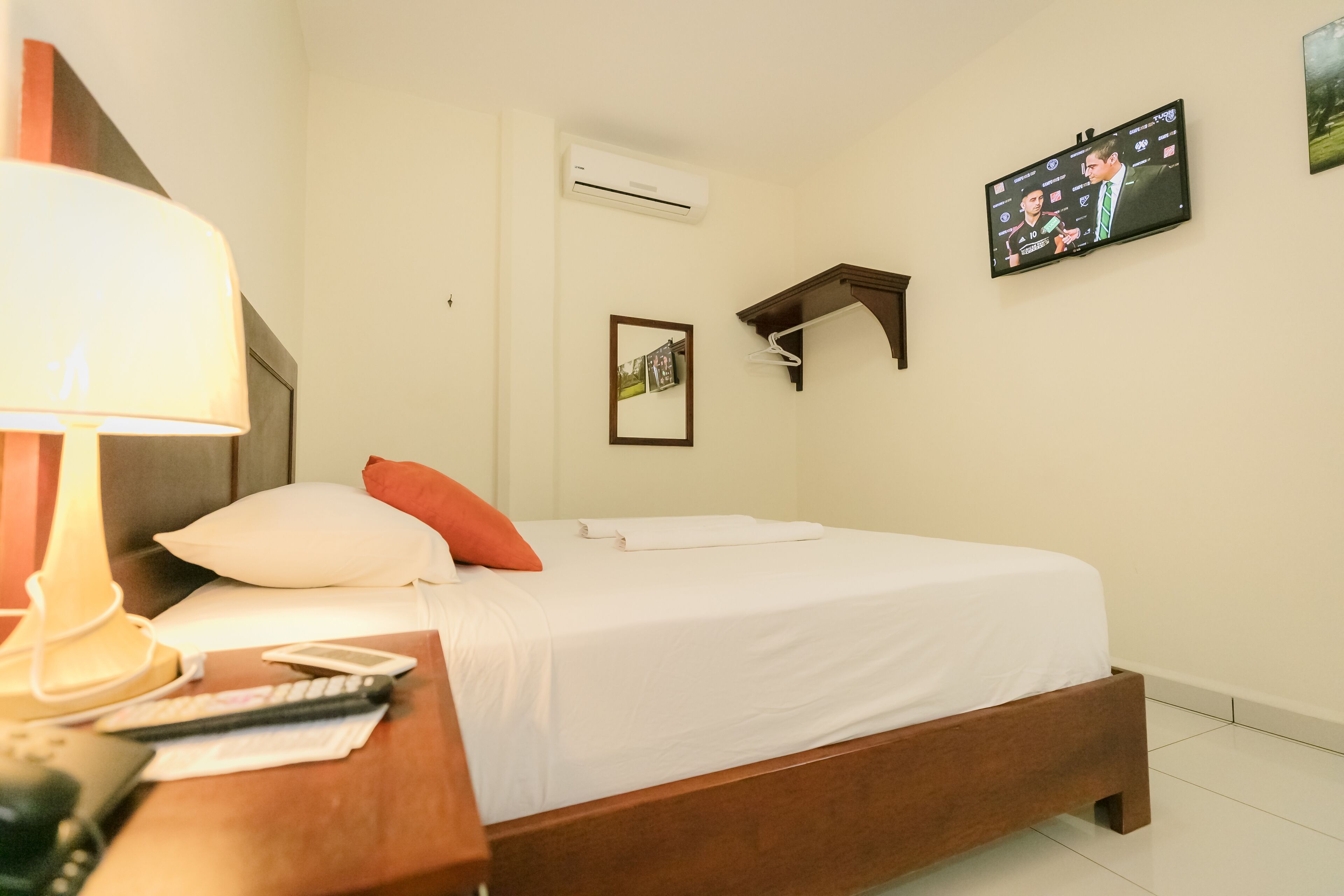 Una habitación de hotel con cama, una mesita de noche con lámpara, un televisor en la pared y un equipo de aire acondicionado.