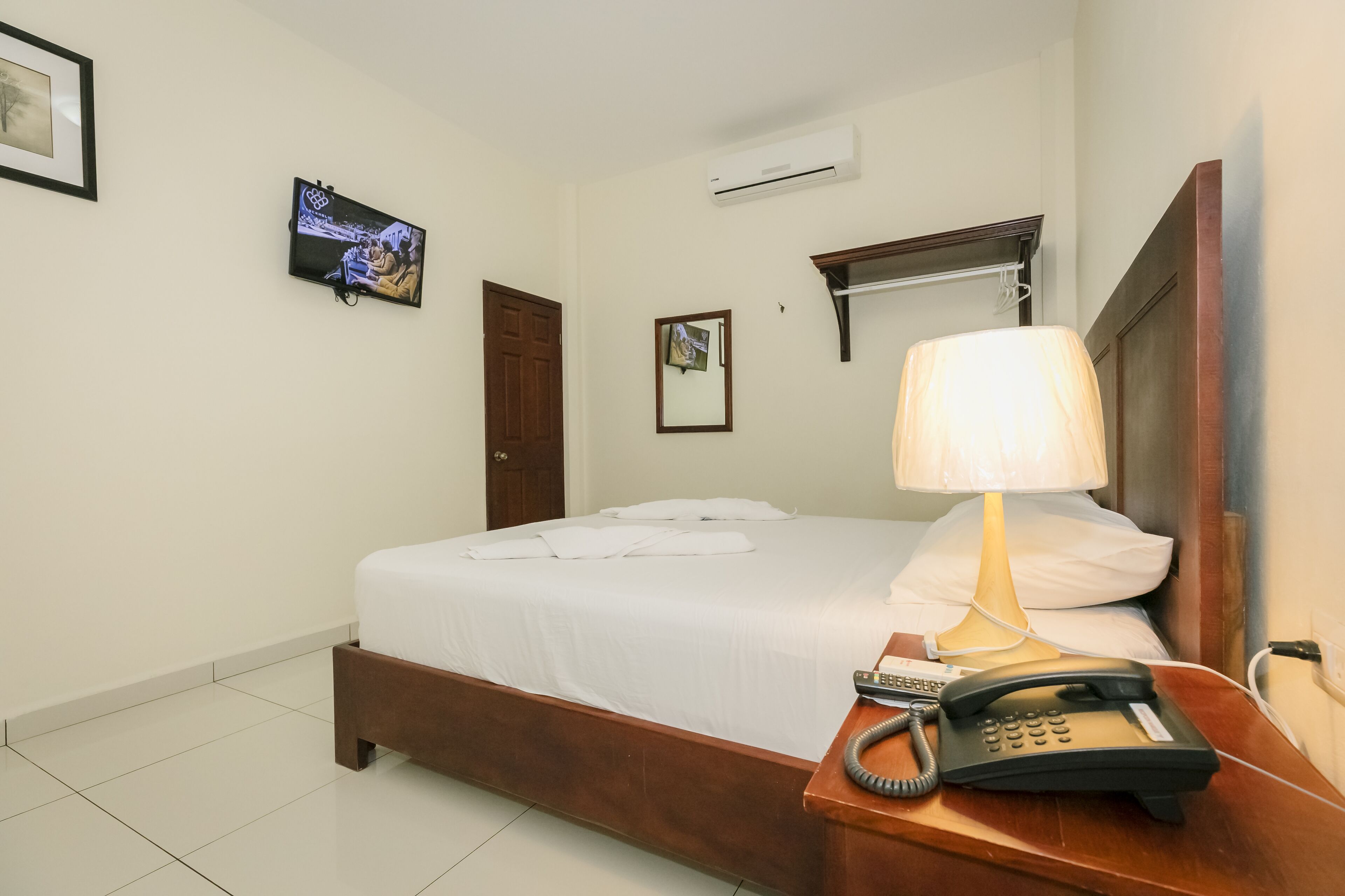 Ein Hotelzimmer mit einem Bett, einer Nachttischlampe, einem Telefon, einer Klimaanlage, einem Fernseher und einem gerahmten Bild an der Wand.