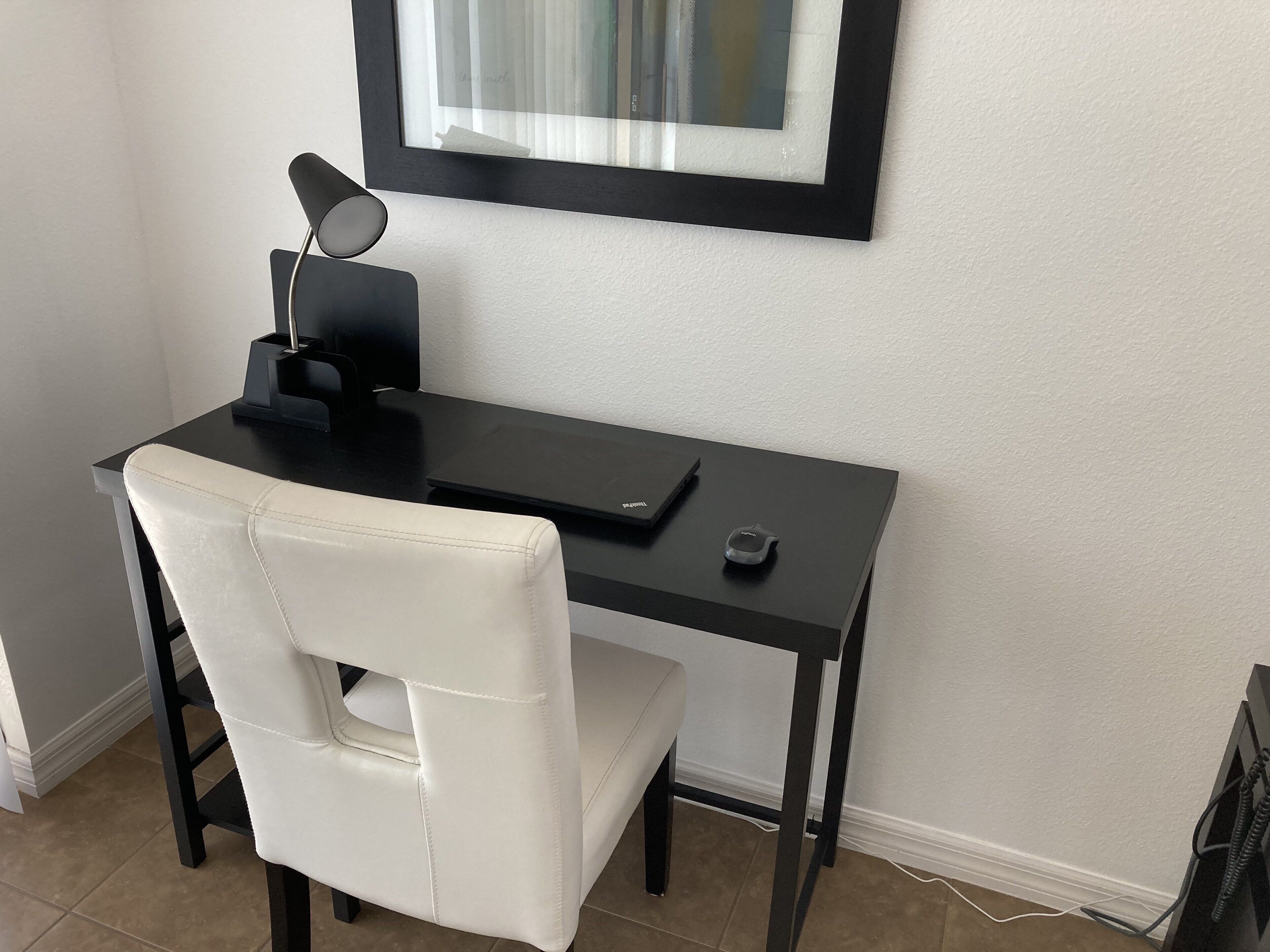 Un petit bureau moderne, doté d’un bureau noir, d’une chaise en cuir blanc, d’un tableau encadré au mur et d’une petite lampe de bureau.