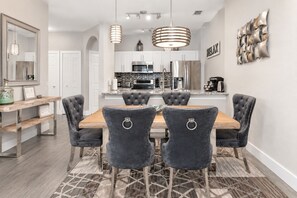 A dining area with a wooden table and four upholstered chairs, a kitchen in the background, and a decorative wall piece.