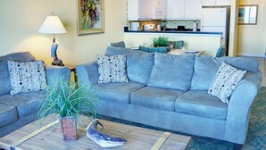 A living room with a grey sofa, patterned cushions, a coffee table with a plant and decorative items, a lamp, and a kitchen in the background. - Ocean Front Two Bedroom Lock Off, Daytona Beach 2056760 (Daytona Beach)