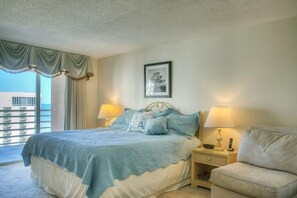 A hotel room with a bed, two bedside lamps, a nightstand, a chair, a framed picture on the wall, and a view of the sea through a window with curtains.