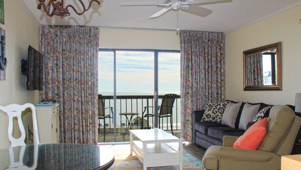 A living room with a glass table, white armchair, and a sofa with patterned cushions. - Ocean Dunes Condo Rental - Right Near the Sands (Myrtle Beach)