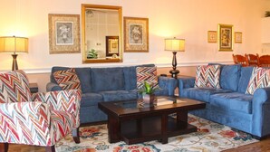 A living room with two blue sofas, a coffee table, and two armchairs with patterned cushions. There are framed pictures on the wall and two lamps on either side of the sofas. - Great Views of the Pool and Grilling Area! (Myrtle Beach)