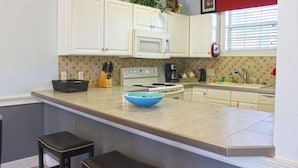 A kitchen with white cabinets, a microwave, a stove, and a sink. There are two stools and a blue bowl on the counter. - Peacefulmyrtlewood Villa Right in the Heart of MB (Myrtle Beach)