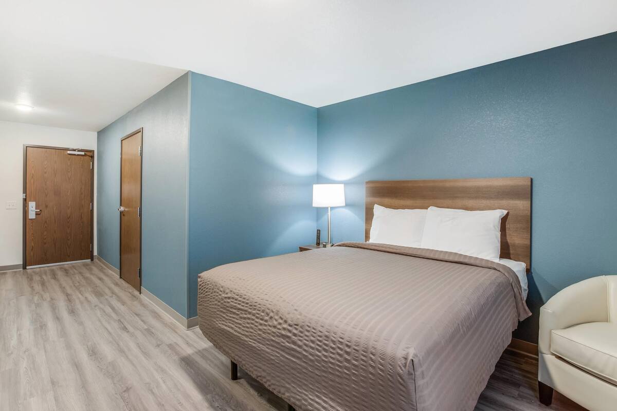 A hotel room with a bed, a nightstand, a lamp, a chair, and a door.