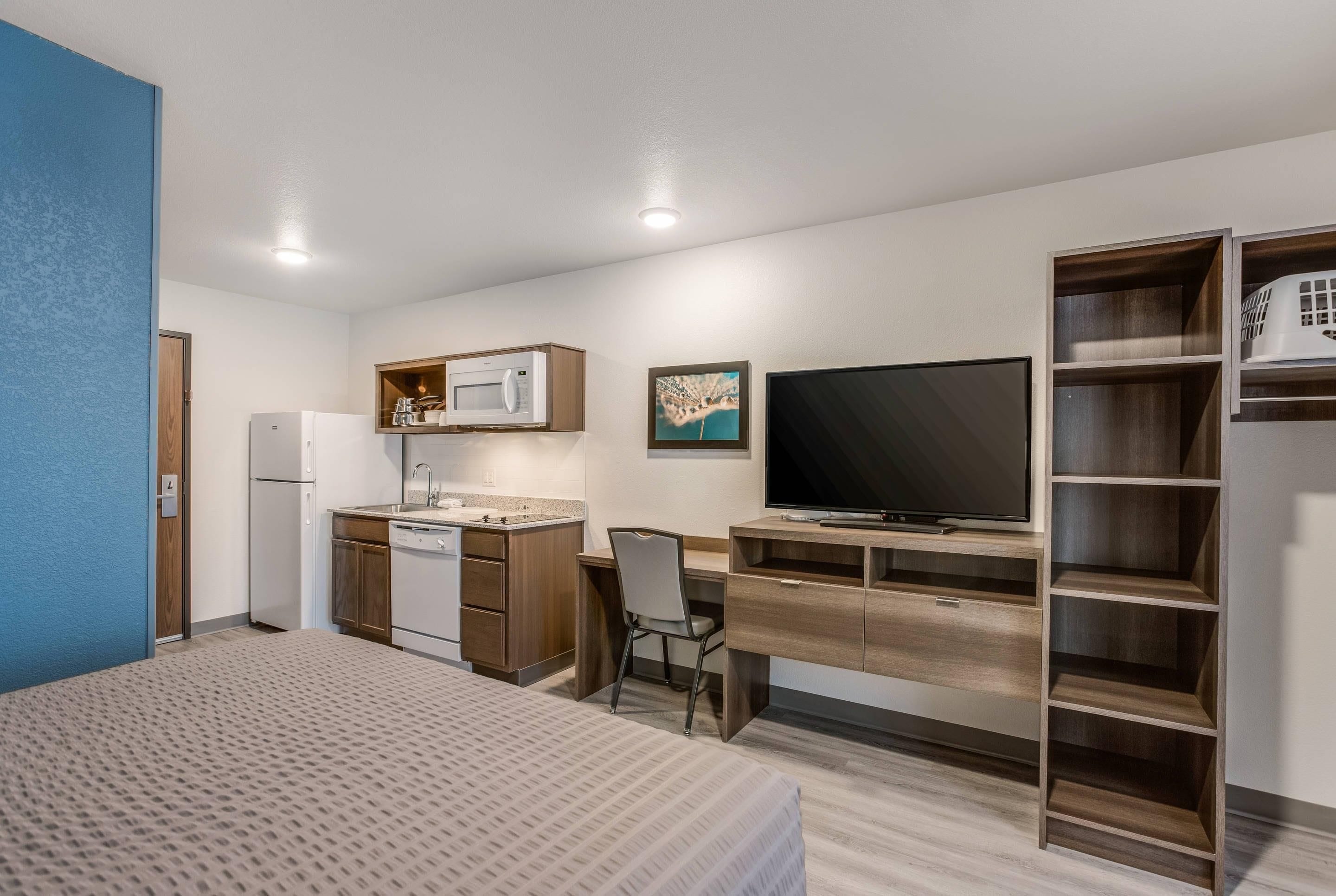 A compact room with a bed, a desk, a chair, a television, a microwave, and a small kitchenette.