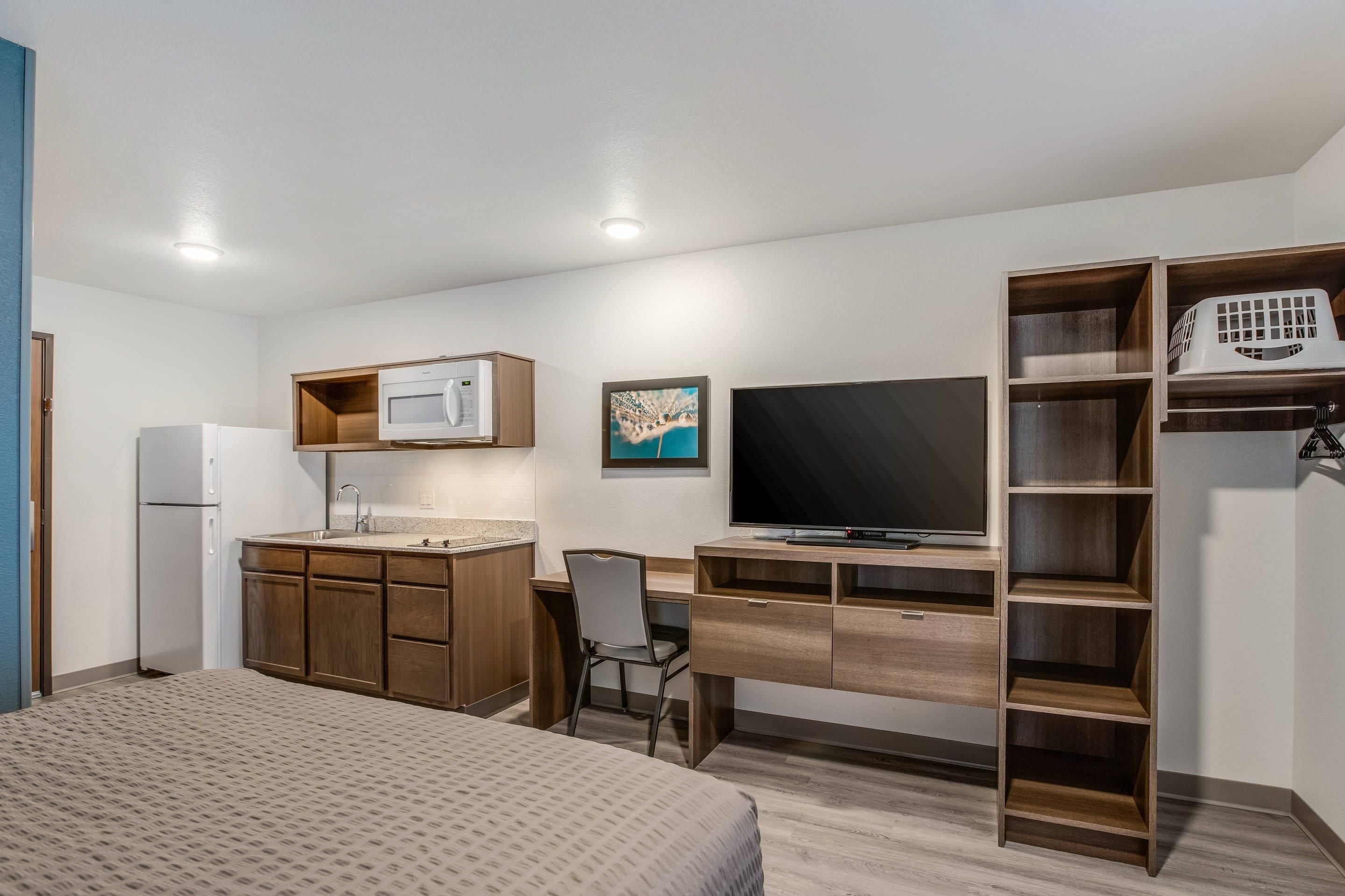 A compact hotel room with a bed, kitchenette, desk, and TV.