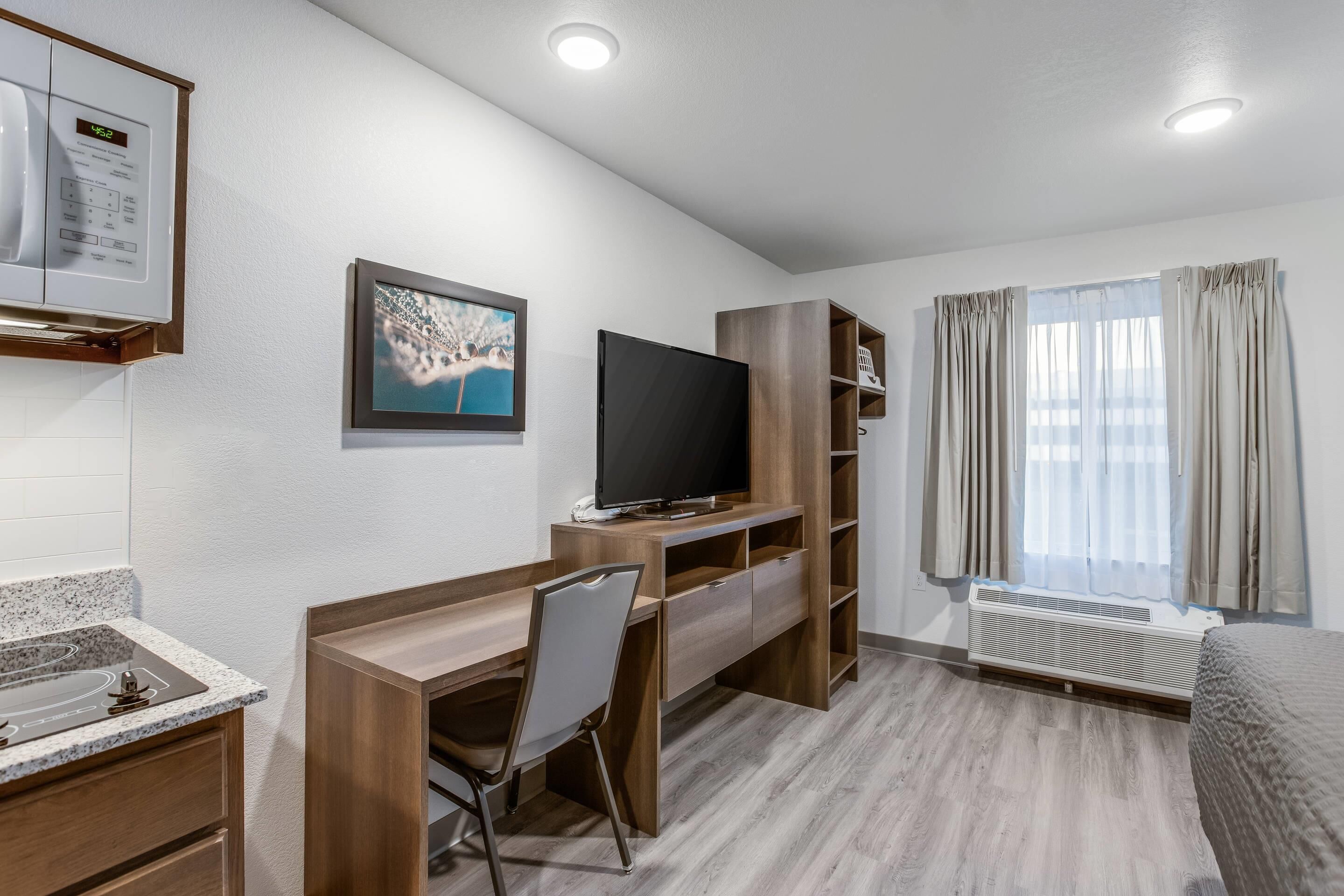 Moderni hotellihuone, jossa on puinen työpöytä, litteänäyttöinen televisio, sisäänrakennettu ilmastointilaite, sänky ja ikkuna, jossa on verhot.